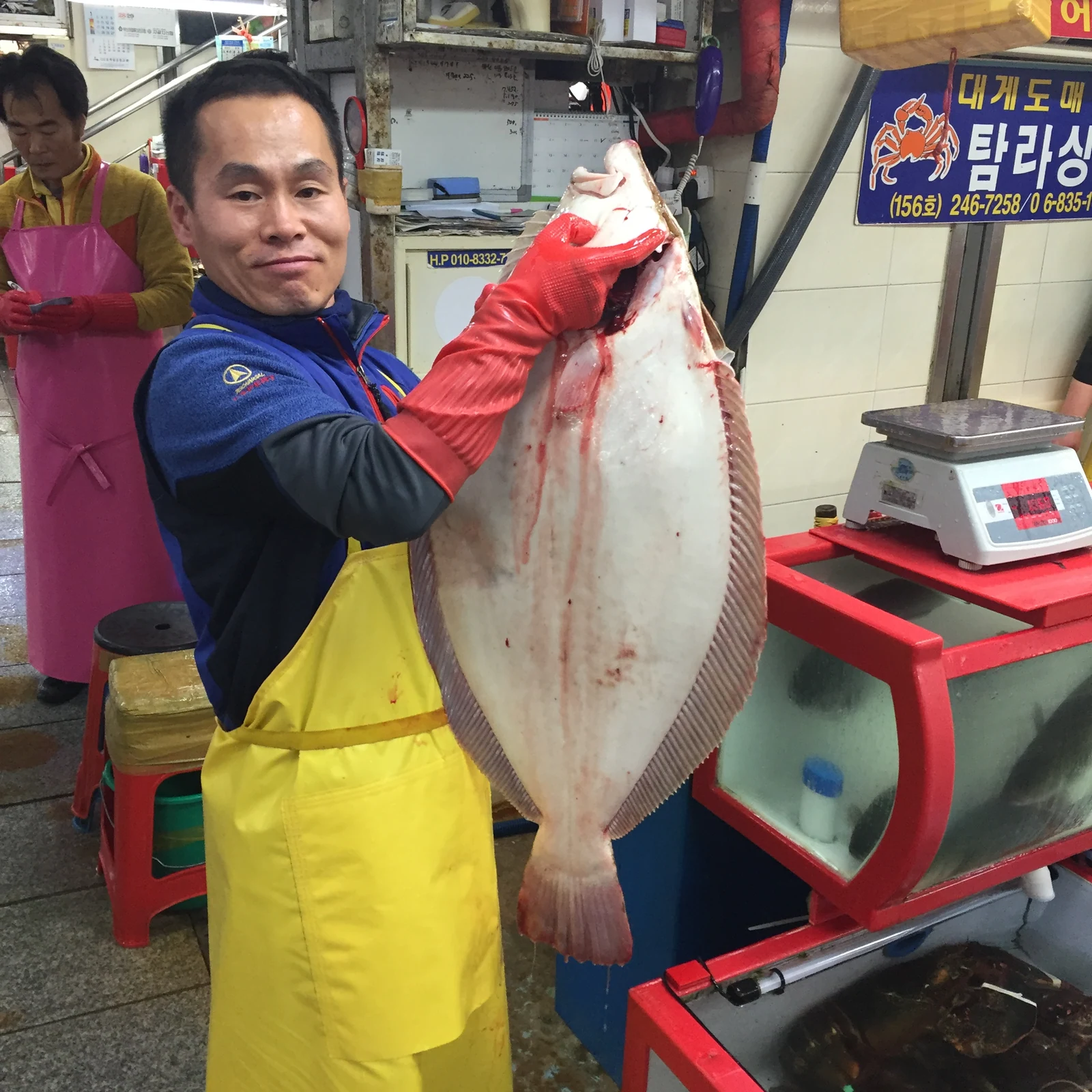 탐라상회 사장이 손질한 대형 광어를 두 손으로 들어 보이는 모습. 뒤로 저울과 붉은 프레임의 활어 수조가 있다.